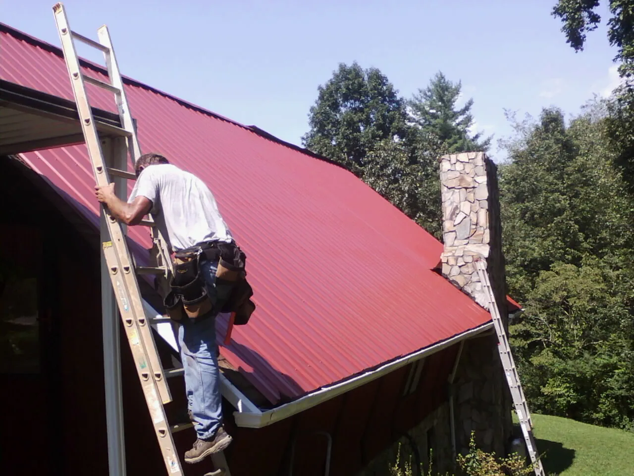 Roofer on a ladder beside a red metal roof and stone chimney — Soffit & Fascia Repair by Roofing Brothers in Parksley
